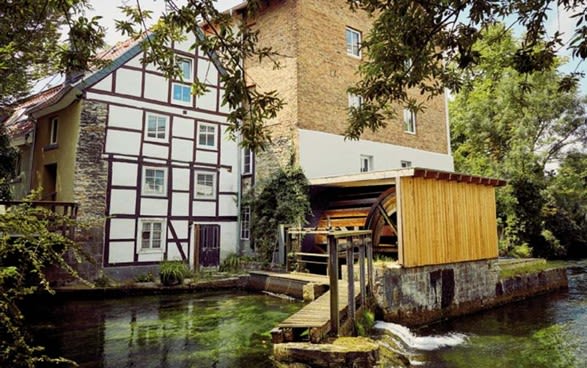 Das Bild zeigt das Wasserrad neben einem Fachwerkhaus, im Vordergrund fließt der Fluss Pader.